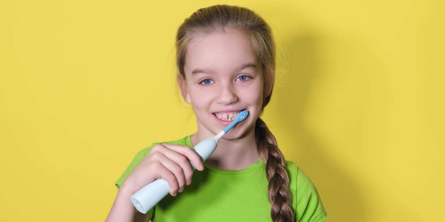 Teenager girl brushes her teeth on yellow background. Child girl in green t-shirt uses an electric toothbrush hoe poets je je tanden?